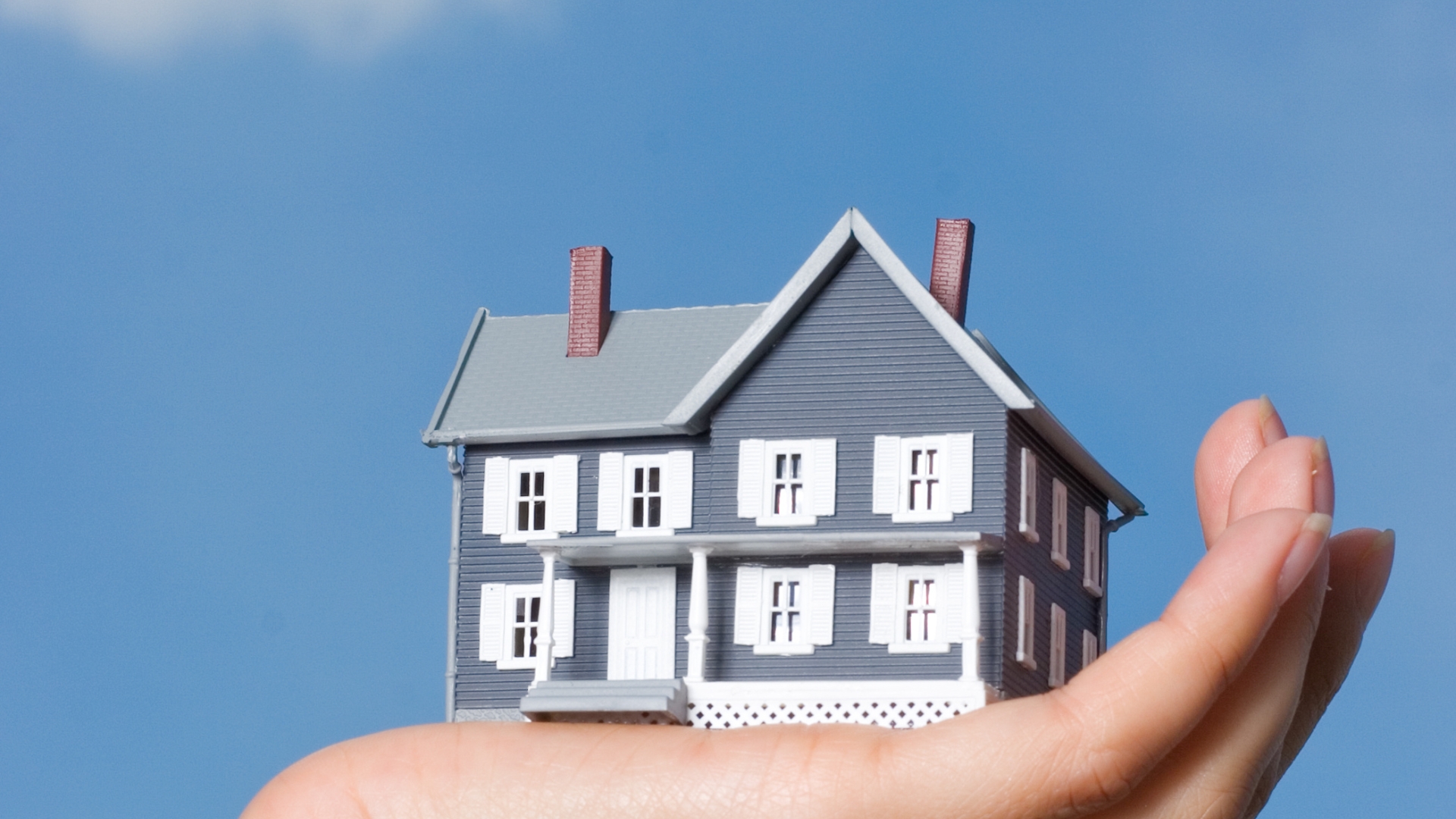 A hand holding a small model house in front of a blue sky